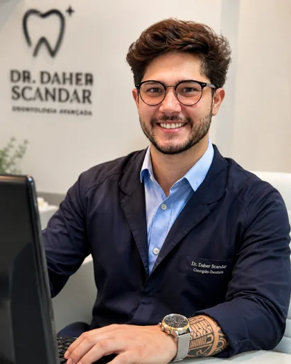 Dr. Daher Scandar sentado à mesa de atendimento, de blazer e óculos.