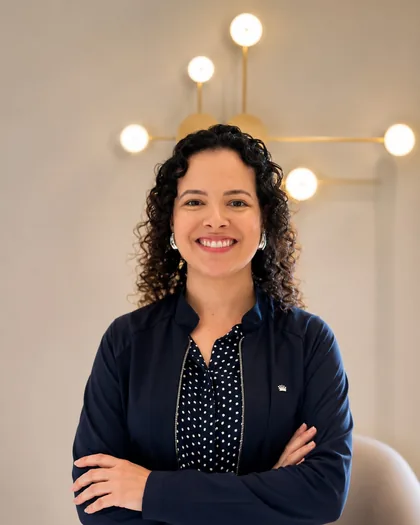 Dra. Thais Cougo Gomes de braços cruzados, em blazer azul-marinho, num ambiente claro da clínica.